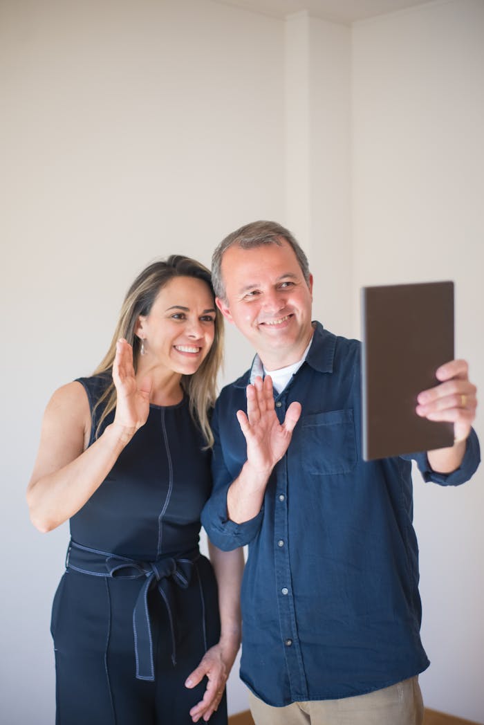 A joyful couple making a videocall from their bright home interior, showing a welcoming gesture.
